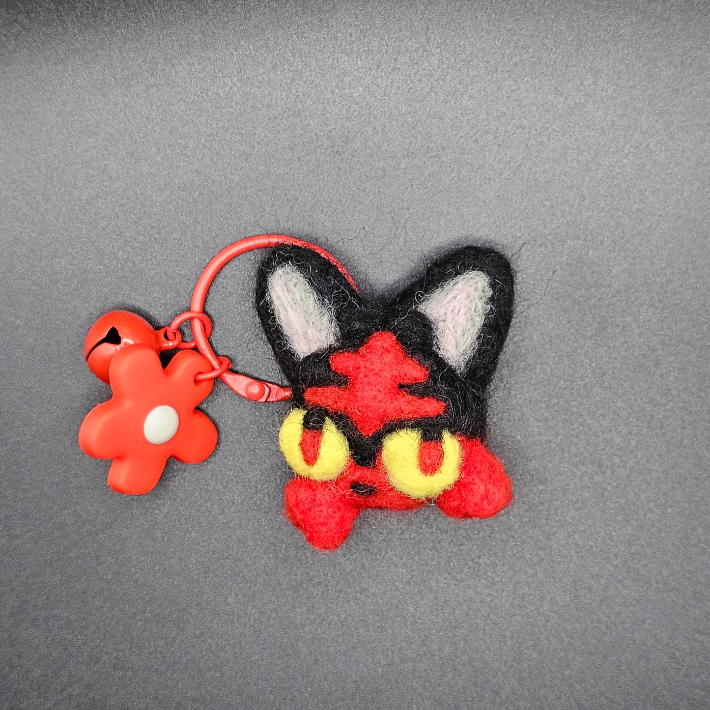 eedle-felted head of black and red fire kitty. Fire kitty has a red pattern on it's forehead and tufts of fur where whiskers would be. The fur tufts appear like flames on the side of the kitty's face. Fire kitty has yellow eyes with black and red pupils. Pictured as keychain with red keyring and red flower charm.