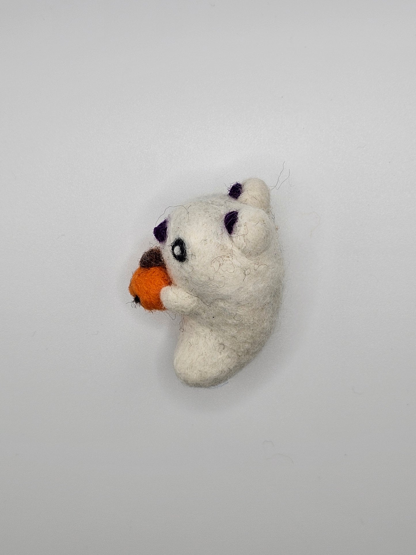 Needle-felted ghost with cat ears and nose. Inside of ears and nose are purple. Ghost cat is holding a tiny Jack-o'-lantern. Profile view of figurine.