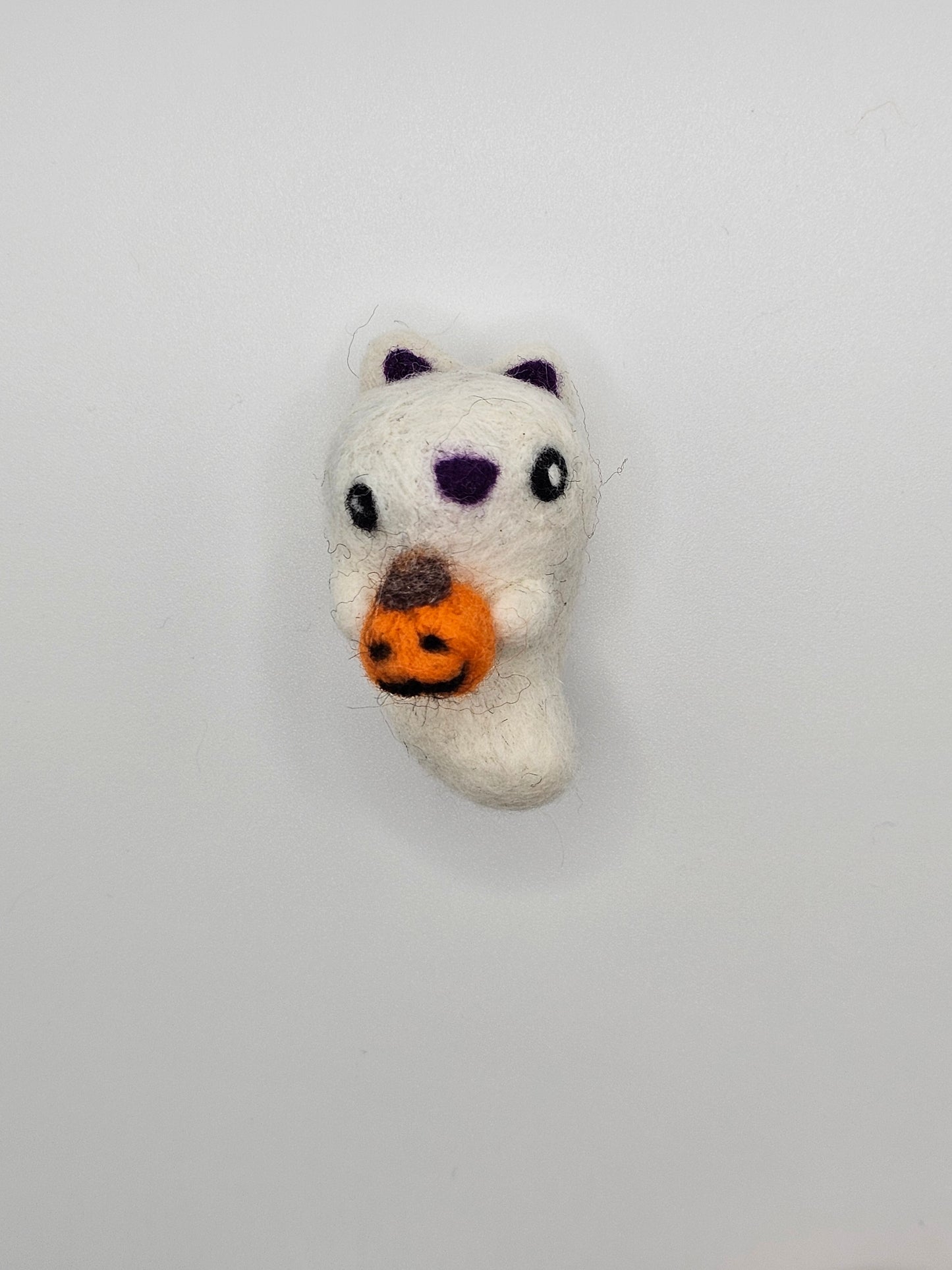 Needle-felted ghost with cat ears and nose. Inside of ears and nose are purple. Ghost cat is holding a tiny Jack-o'-lantern.