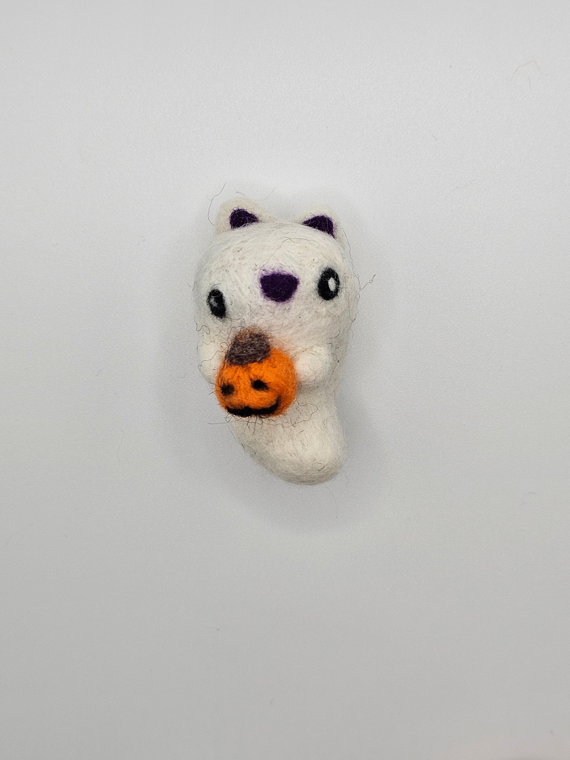 Needle-felted ghost with cat ears and nose. Inside of ears and nose are purple. Ghost cat is holding a tiny Jack-o'-lantern.