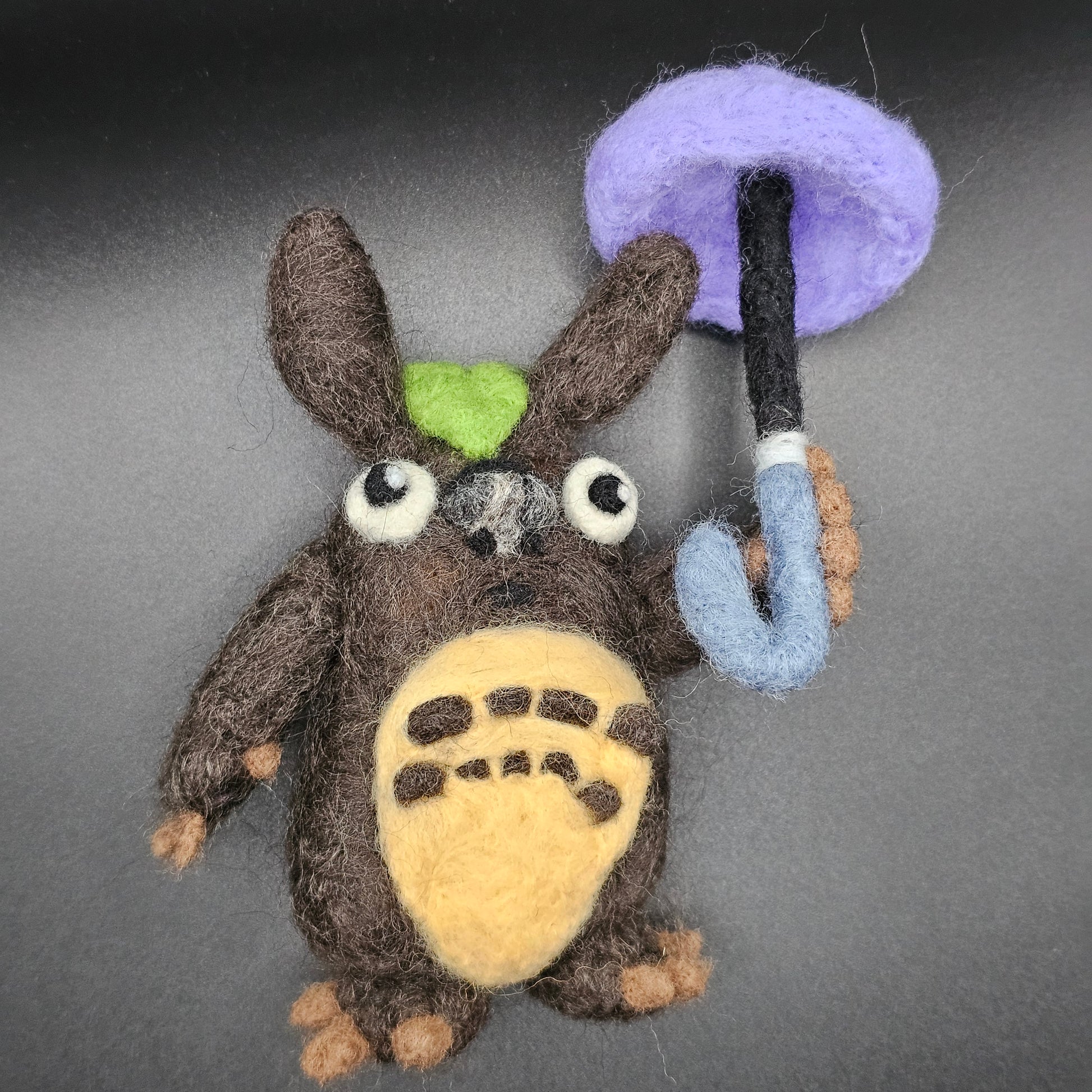 Needle-felted figurine inspired by Totoro. Totoro is a blue cat-bear-owl hybrid. This interpretation is brown, has a cabbage leaf on his head, and holds a purple umbrella.