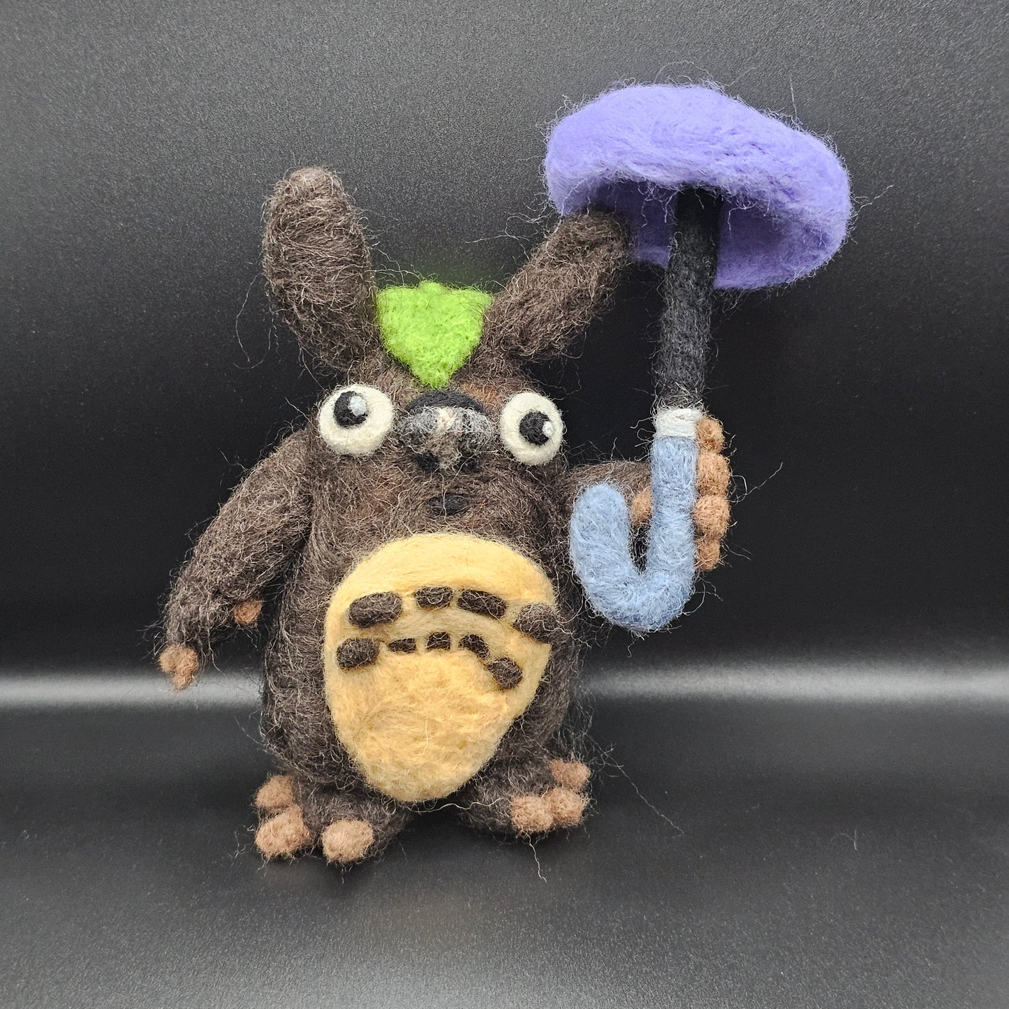 Needle-felted figurine inspired by Totoro. Totoro is a blue cat-bear-owl hybrid. This interpretation is brown, has a cabbage leaf on his head, and holds a purple umbrella.