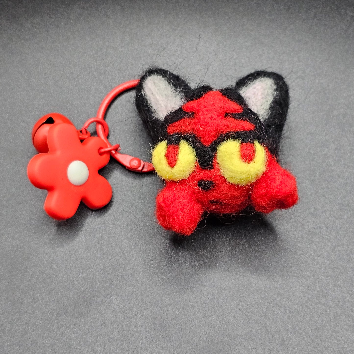 eedle-felted head of black and red fire kitty. Fire kitty has a red pattern on it's forehead and tufts of fur where whiskers would be. The fur tufts appear like flames on the side of the kitty's face. Fire kitty has yellow eyes with black and red pupils. Pictured as keychain with red keyring and red flower charm.