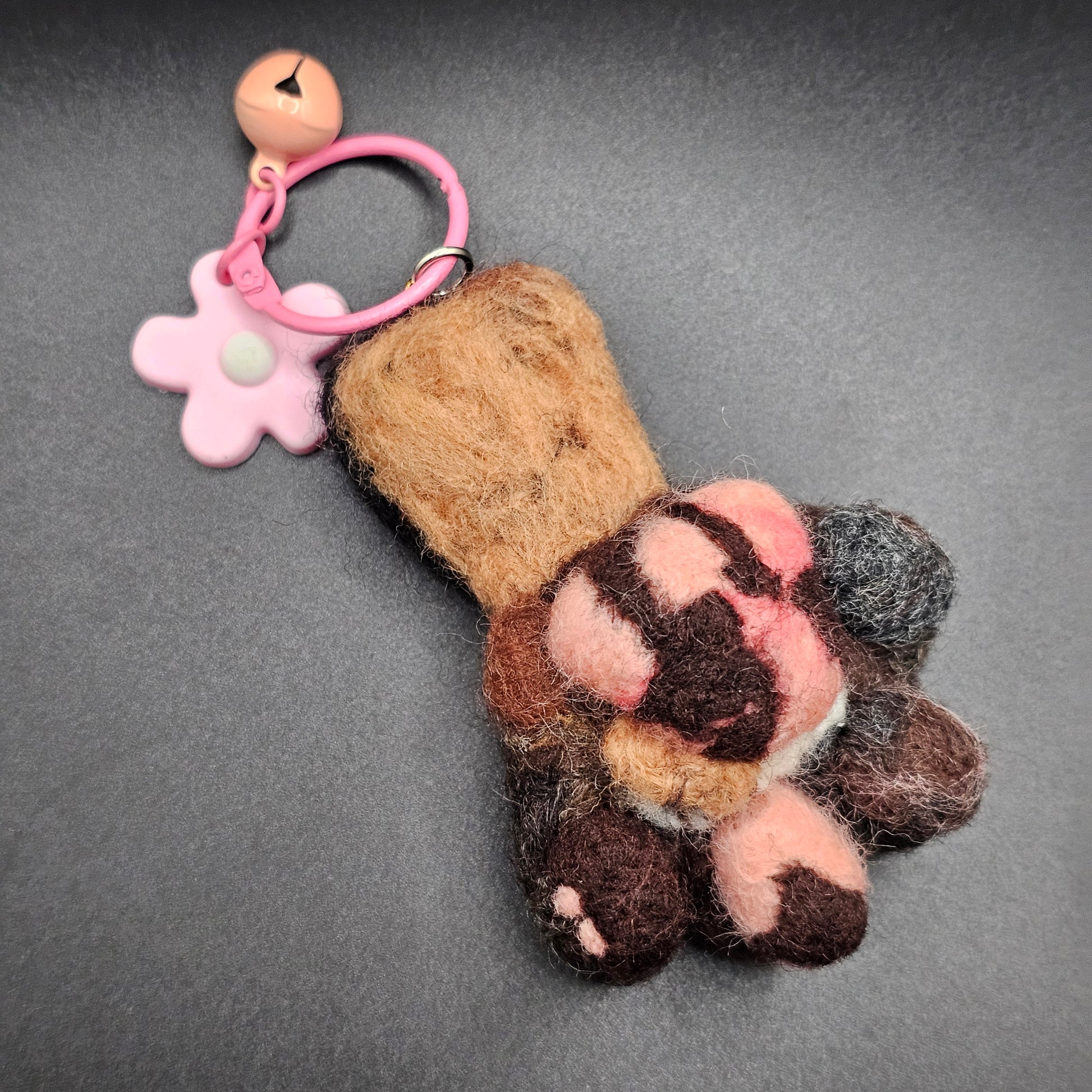 Needle-felted Tortoise Shell cat paw. Toe beans are brown, pink with a brown freckle, brown, and black. Paw pad is pink with another shade of pink, and brown freckles. Pictured aa a keychain with a pink keyring and pink flower charm.