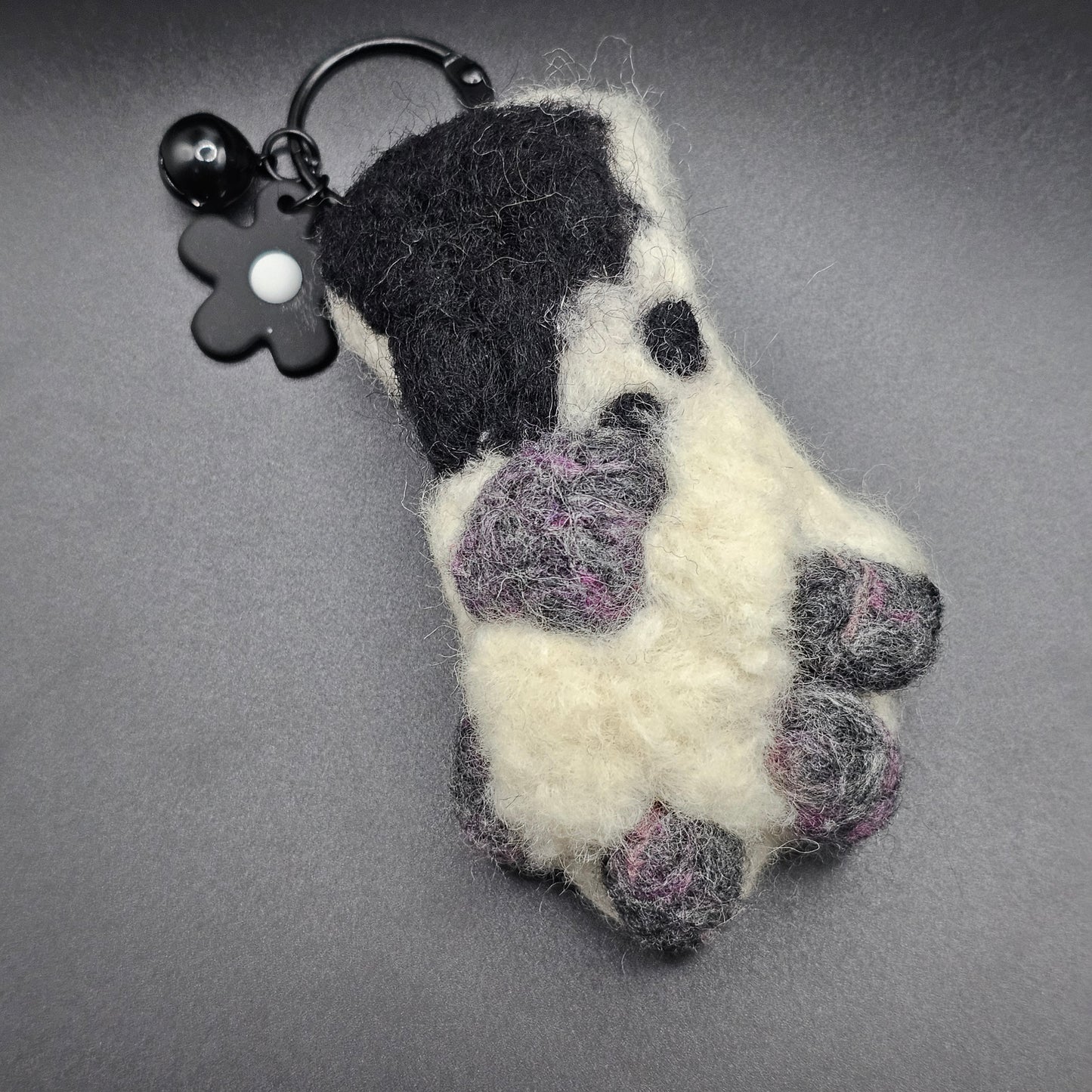 White fluffy paw with grey and pink speckled toe beans and paw pad. Ankle is a lighter grey with a large black spot. Pictured as keychain with black keyring and flower charm.