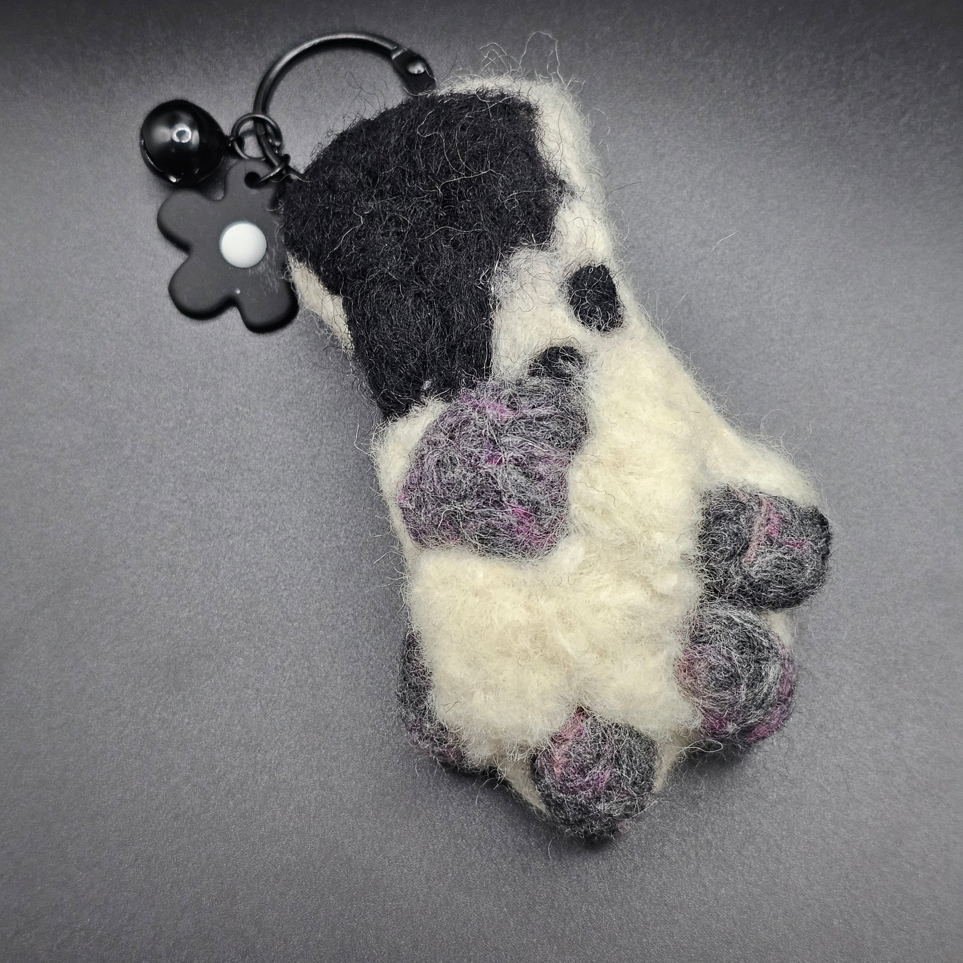 White fluffy paw with grey and pink speckled toe beans and paw pad. Ankle is a lighter grey with a large black spot. Pictured as keychain with black keyring and flower charm.