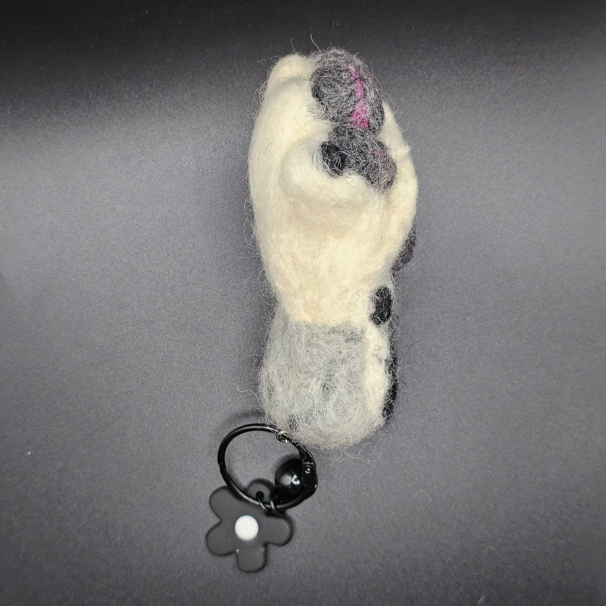 White fluffy paw with grey and pink speckled toe beans and paw pad. Ankle is a lighter grey with a large black spot. Pictured as keychain with black keyring and flower charm. Profile view.