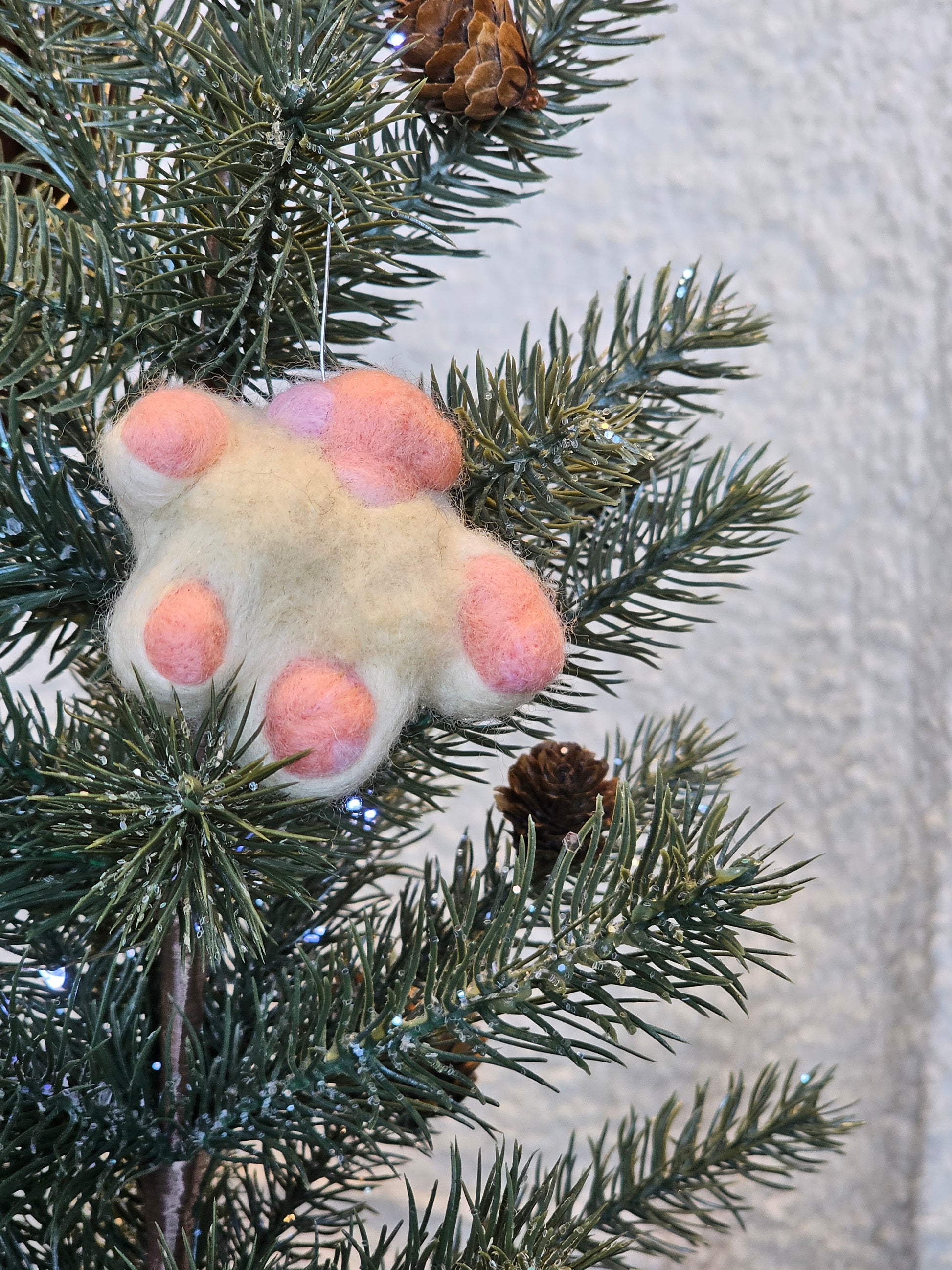 Needle-felted white cat paw with pink toe beans and pink paw pad. Pictured as ornament in a tree.