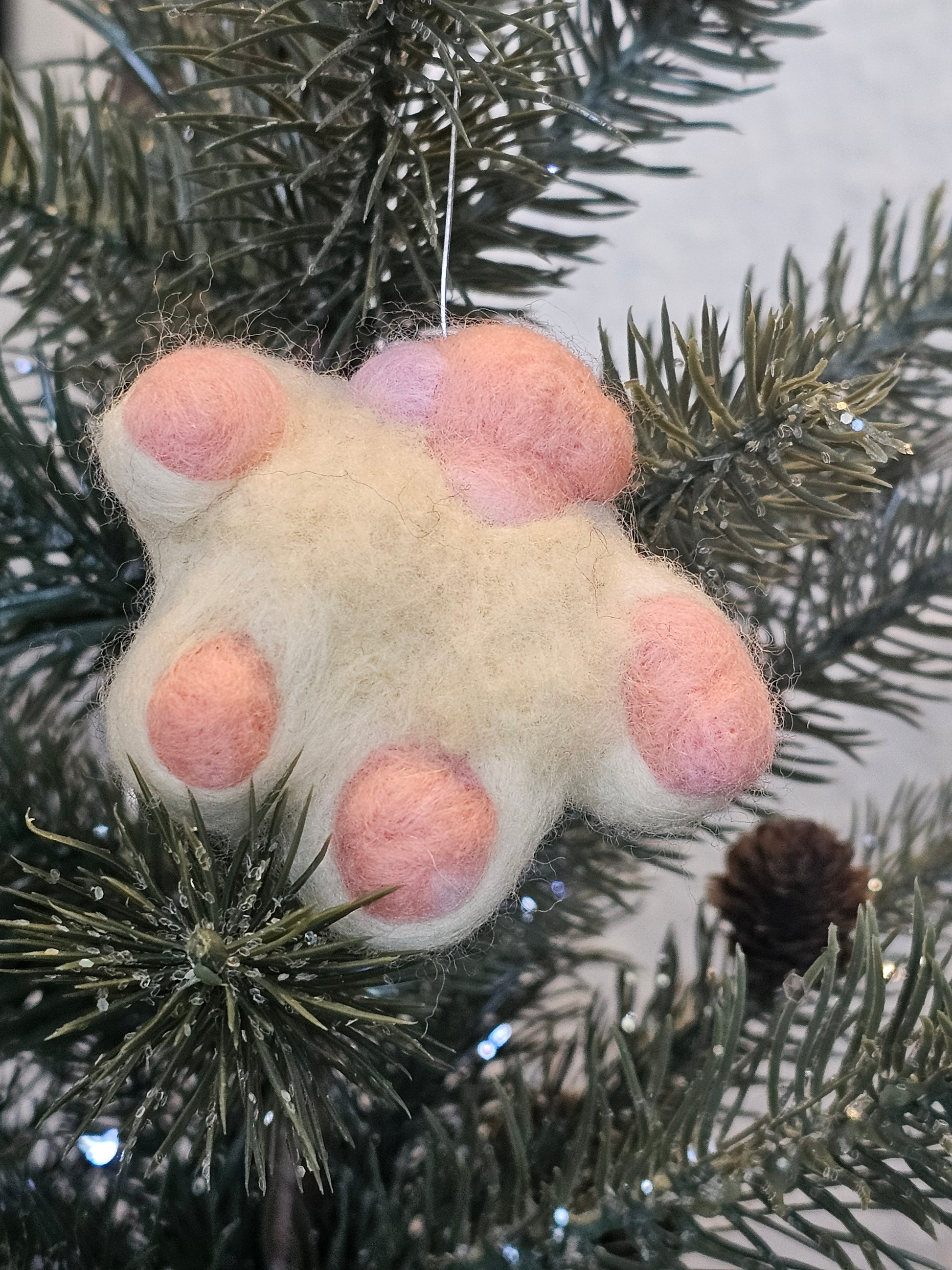 Needle-felted white cat paw with pink toe beans and pink paw pad. Pictured as ornament in a tree.