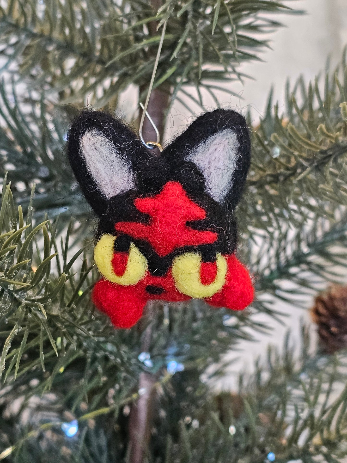 Needle-felted head of black and red fire kitty. Fire kitty has a red pattern on it's forehead and tufts of fur where whiskers would be. The fur tufts appear like flames on the side of the kitty's face. Fire kitty has yellow eyes with black and red pupils. Pictured hanging in Christmas tree 