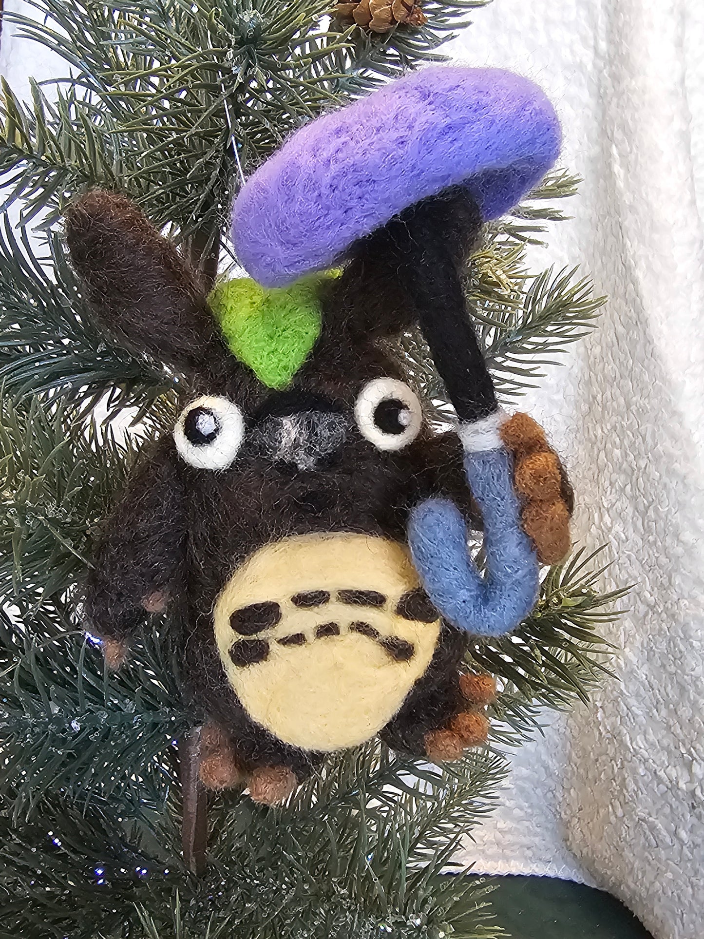 Needle-felted figurine inspired by Totoro. Totoro is a blue cat-bear-owl hybrid. This interpretation is brown, has a cabbage leaf on his head, and holds a purple umbrella. Pictured hanging in a Christmas tree.
