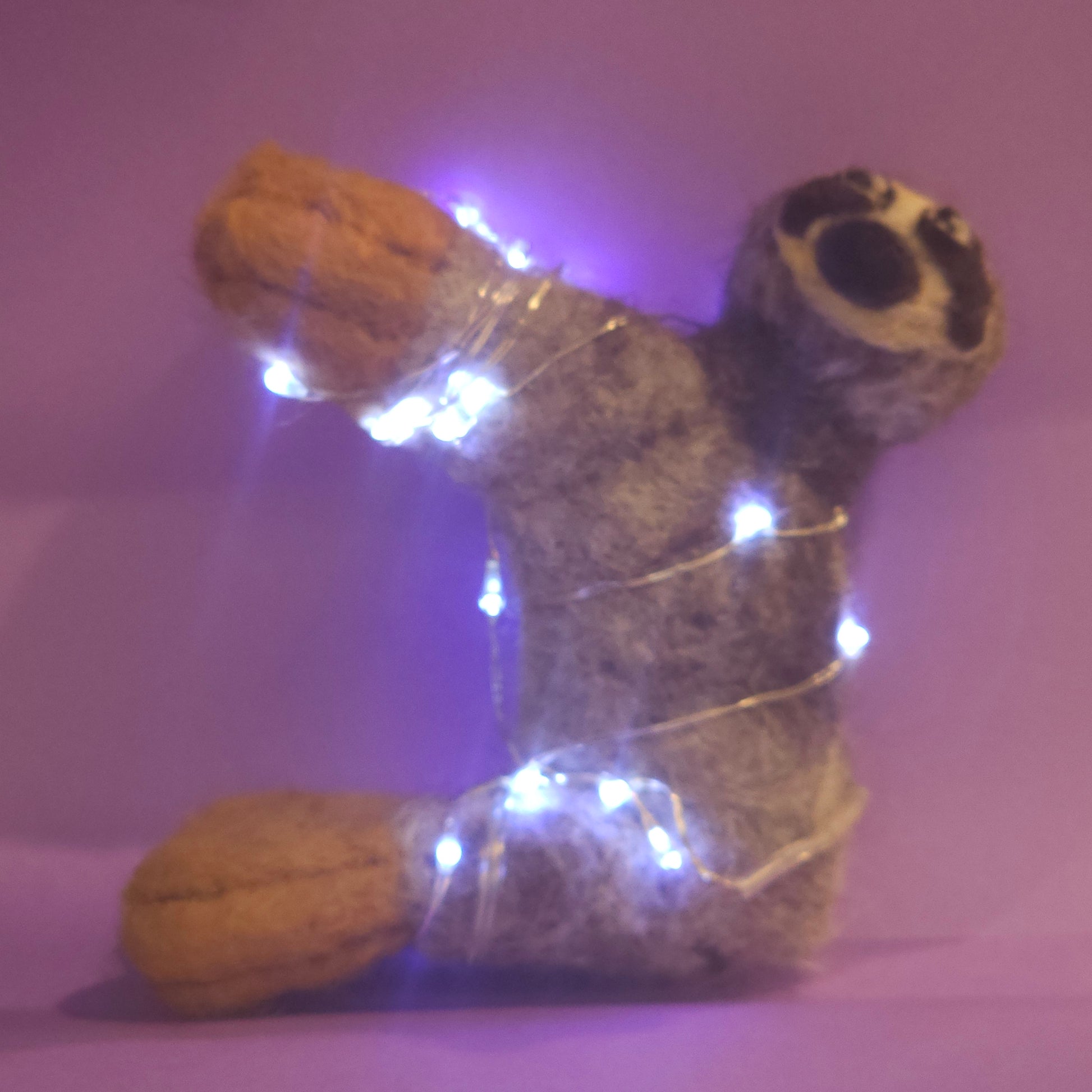 Needle-felted sloth sitting upright in front of a purple backdrop. The sloth is wrapped in twinkle lights; the twinkle lights are shining blue.
