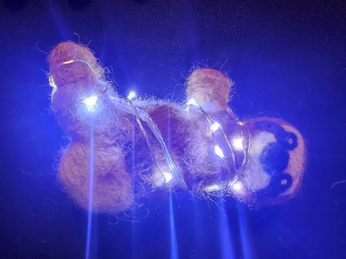 Needle-felted sloth laying on back, wrapped in twinkle lights. The twinkle lights are shining with a blue hue.