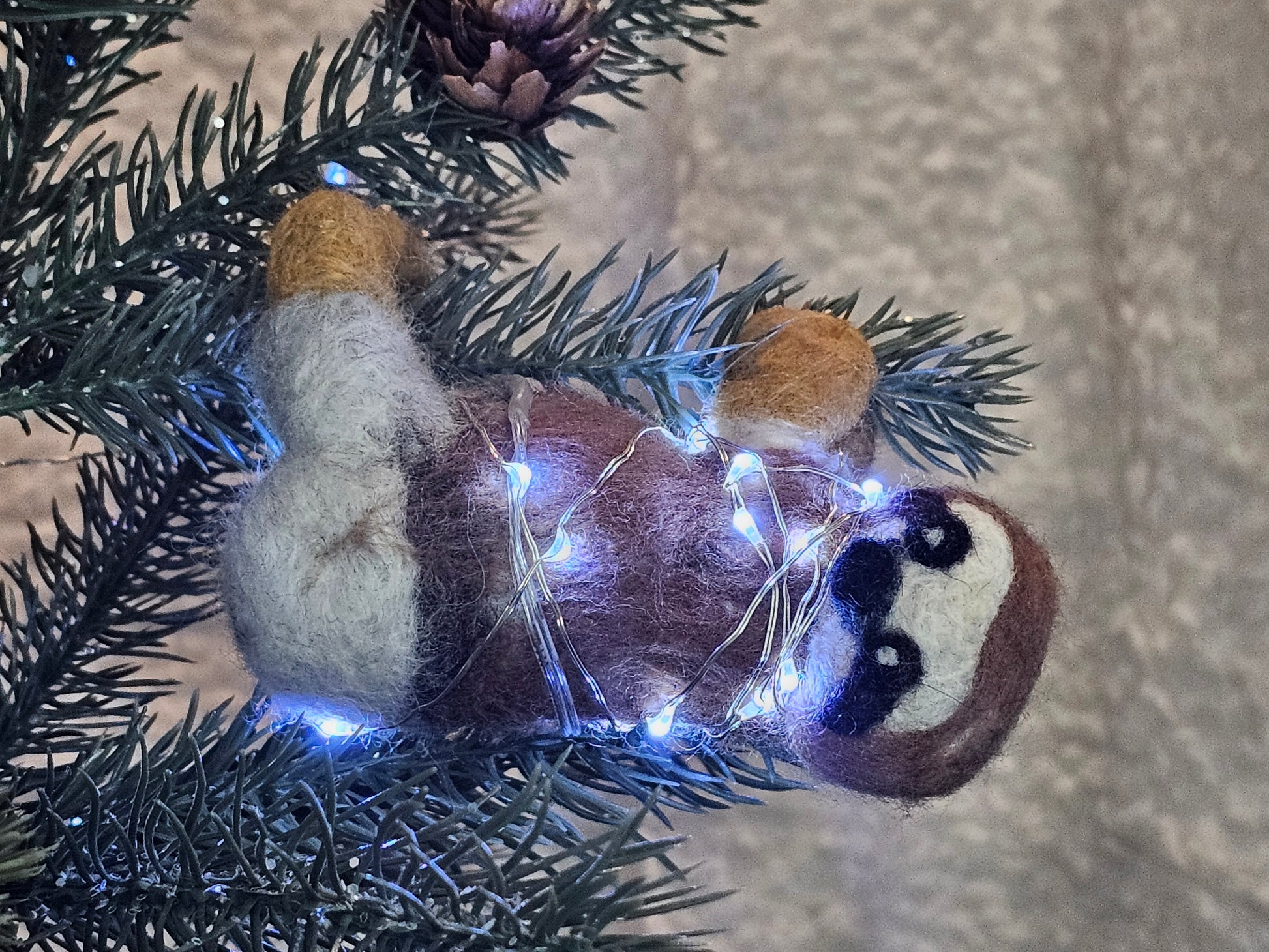 Needle-felted sloth hanging onto tree branch with all four legs, with twinkle lights lit up.