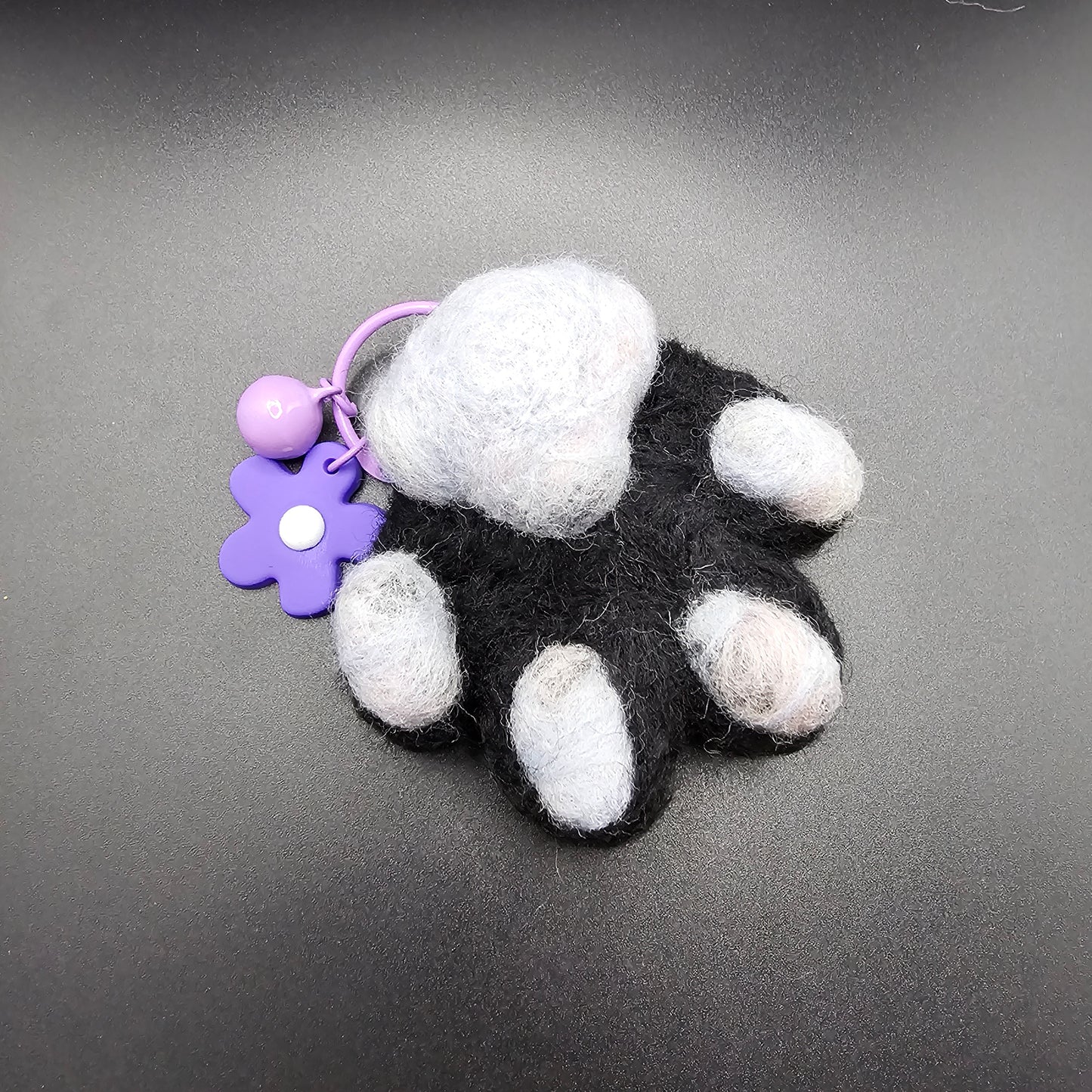 Needle-felted black cat paw with grey toe beans and a grey paw pad. Has a purple keyring with purple flower charm.